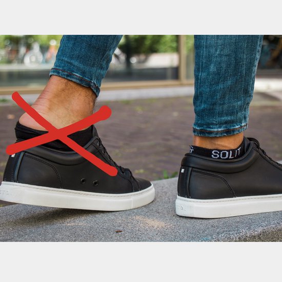 Zwarte lage sokken met witte 'SOLIT' opdruk, gedragen onder jeans en zwarte sneakers. Rode kruis door de rechter enkel.