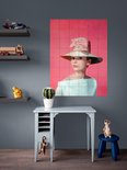 Audrey Hepburn portant un grand chapeau de paille avec un nœud rose, une robe bleu pâle épaules dénudées, sur fond rose, près d'un bureau.