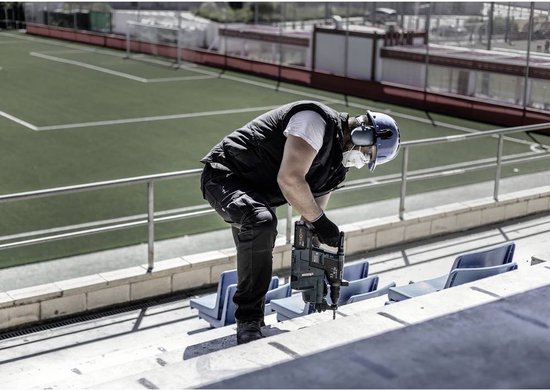 Man met een blauwe helm en gehoorbescherming gebruikt een Bosch boorhamer bij een stadion.