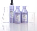 Trois bouteilles Redken avec de la verrerie de laboratoire. Shampooing, après-shampooing et pré-traitement dans des emballages violet clair.