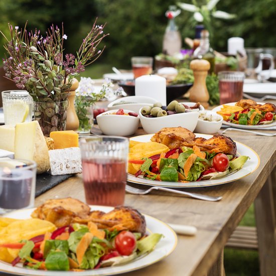Buiten gedekte tafel met witte borden met gouden rand, gevuld met kip, salade en aardappelen, kaas en olijven.