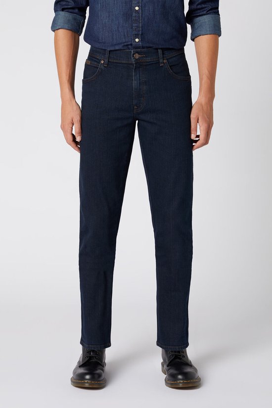 Model draagt een donkerblauwe Wrangler regular fit jeans en een spijkerhemd met zwarte schoenen.