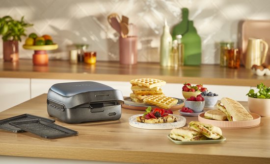 Een donkergrijs Tefal wafelijzer staat op een houten aanrecht, met wafels en panini ernaast.