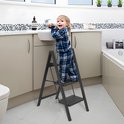 Een kind in pyjama staat op een zwarte 3-treden trap in een badkamer.