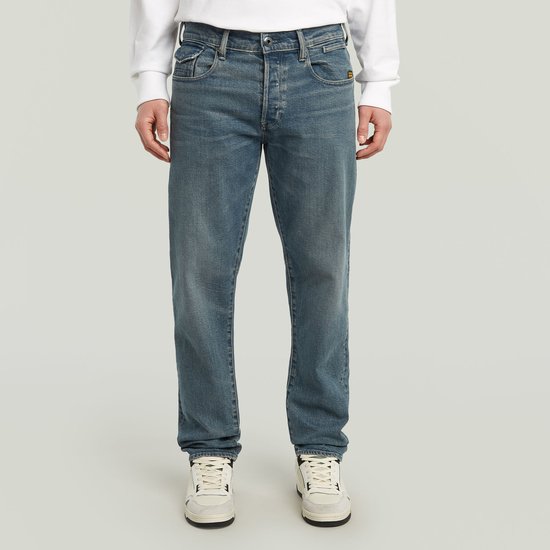 Man met een witte trui en een blauwe jeans. De jeans heeft een normale pasvorm en taps toelopende pijpen.