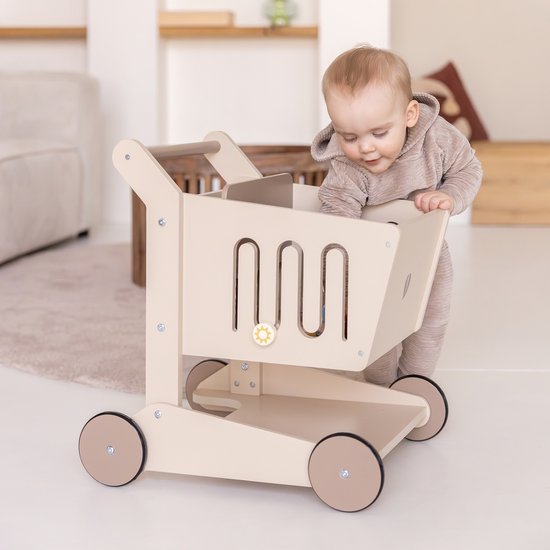 Een baby in een beige outfit leunt over een zandkleurige houten winkelwagen.