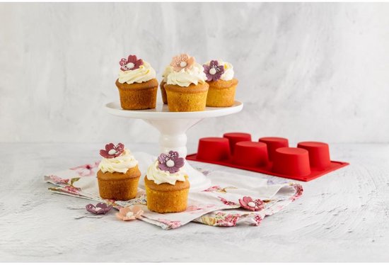 Zes cupcakes met witte crème en bloemen op een witte taartschaal en op een bloemetjesdoek, naast rode muffinvormen.