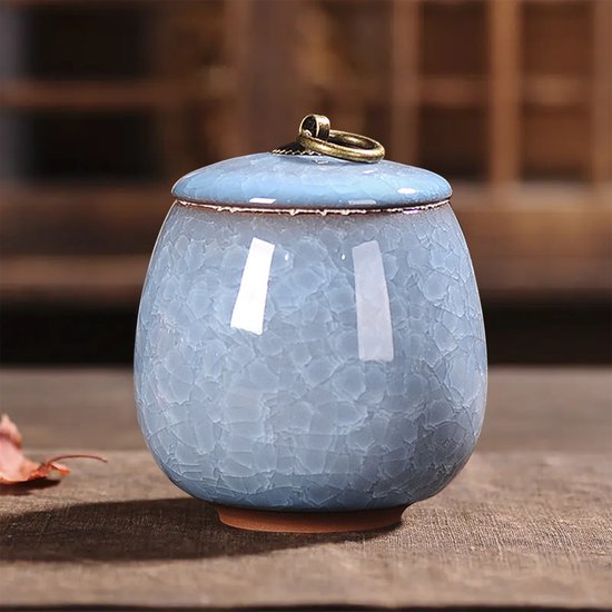Hemelsblauwe keramische pot met deksel, krakelglazuur en gouden ring, staand op een houten tafel.