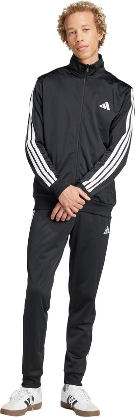 Man in zwart trainingspak met witte strepen, staand, met witte sneakers.