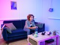 Vrouw met krullend haar lacht terwijl ze een gamecontroller vasthoudt in een woonkamer met roze en blauwe neonverlichting.