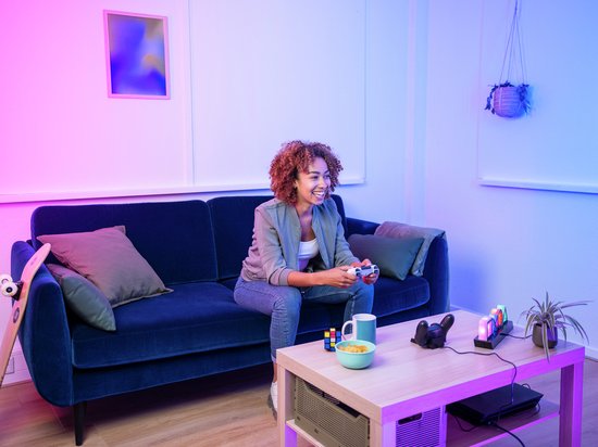 Vrouw met krullend haar lacht terwijl ze een gamecontroller vasthoudt in een woonkamer met roze en blauwe neonverlichting.