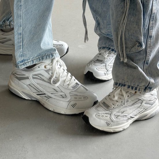 Twee personen dragen witte "DUTCH'D" sneakers met zilveren details en jeans.