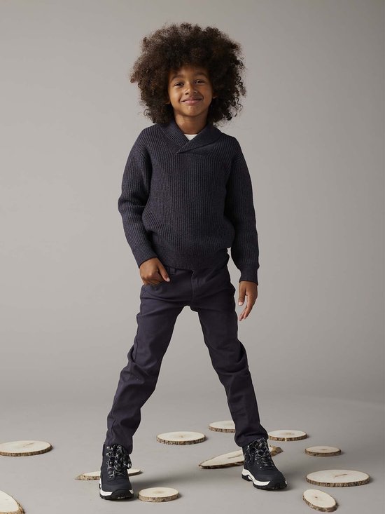 Jongetje met krullend haar in donkerblauw gebreid vest met V-hals en donkerblauwe broek.