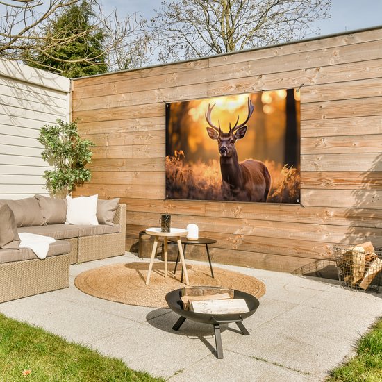 Tuinposter met hert hangt aan een houten muur op een terras met lounge set en vuurkorf.