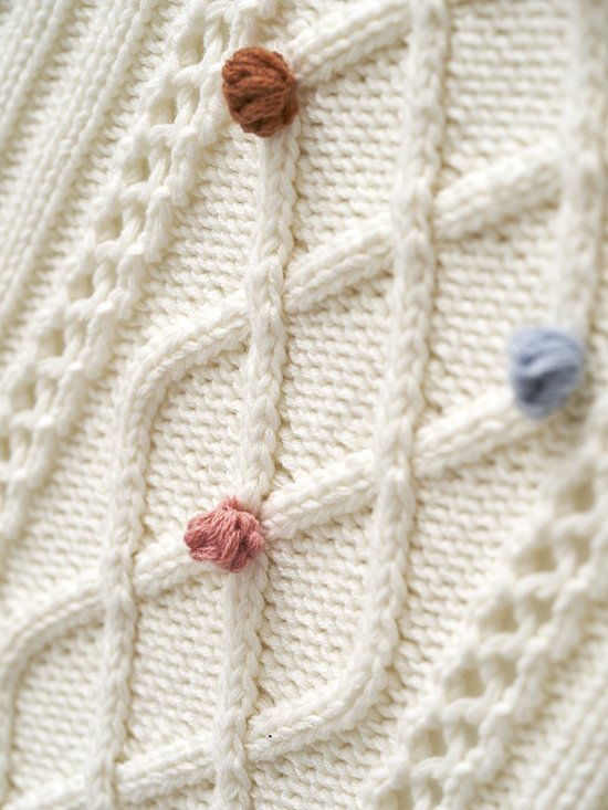 Close-up van off-white gebreide deken met kabels en roze, bruine en blauwe pompons.