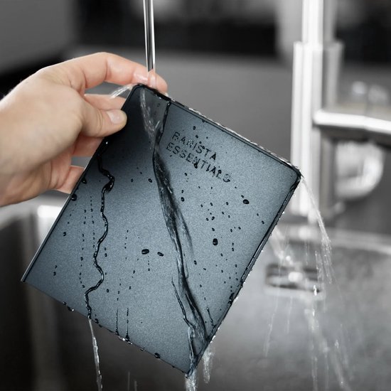 Een zwarte tampermat met de tekst 'BARISTA ESSENTIALS' erop, wordt schoongemaakt onder water.