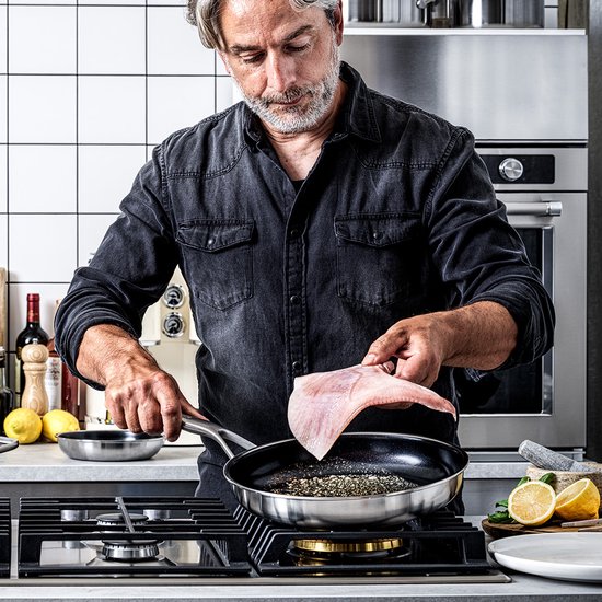 Een man met grijze baard en zwart overhemd bakt een vis in een KitchenAid koekenpan met citroenplakjes.