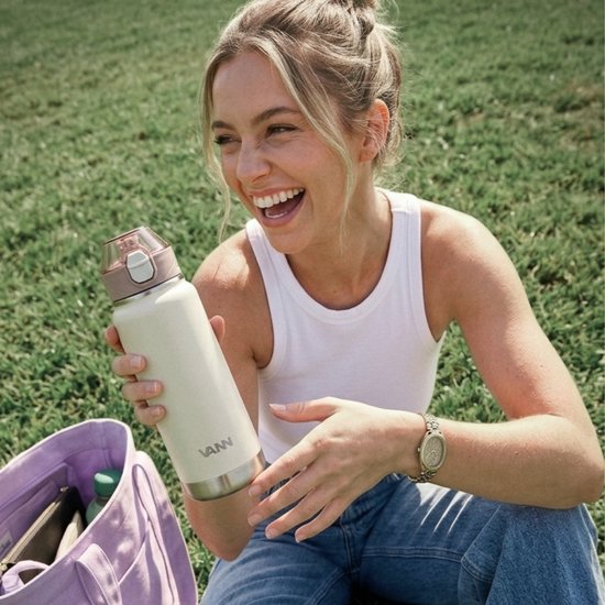 Crème VANN drinkfles met zilveren onderkant, vastgehouden door een lachende vrouw in het gras.
