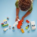 Oranje LEGO Duplo auto chassis met witte bovenkant, omringd door diverse bouwblokken, figuurtjes en een klein geel huisje.