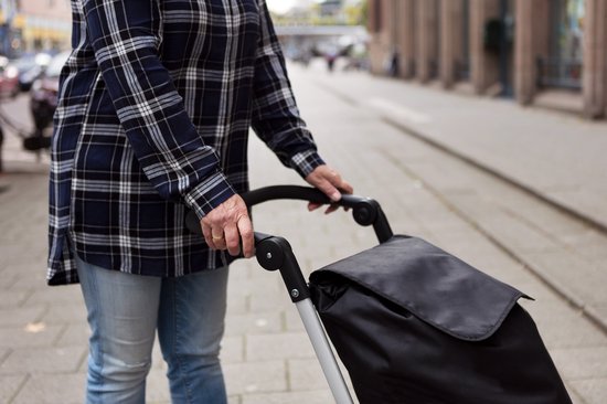 Vrouw met geblokt overhemd en spijkerbroek duwt een zwarte boodschappentrolley op straat.
