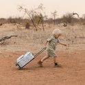 Jongetje met blonde haren in safari-kleding trekt een koffer met safari print door droog gras.