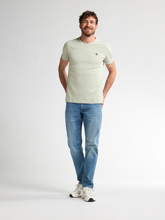 Man glimlacht, draagt lichtgroen T-shirt en lichtblauwe jeans met witte sneakers.