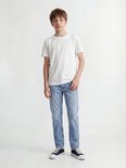 Jongen in wit T-shirt en lichtblauwe jeans, staand, van voren gezien. Draagt sneakers.