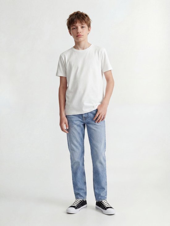 Jongen in wit T-shirt en lichtblauwe jeans, staand, van voren gezien. Draagt sneakers.
