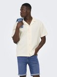 Man met korte mouwen, crèmekleurig hemd met open kraag en knopen. Blauwe jeans shorts en spijkerjasje over de schouder.