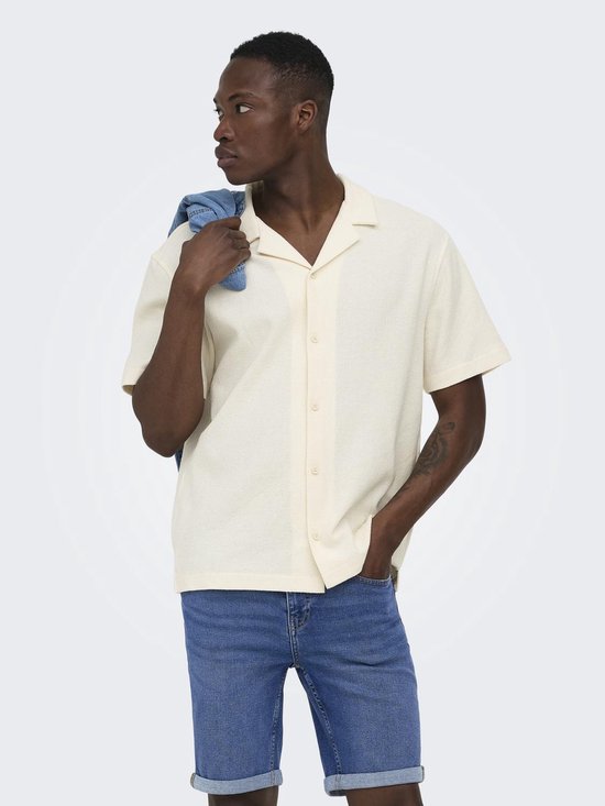 Man met korte mouwen, crèmekleurig hemd met open kraag en knopen. Blauwe jeans shorts en spijkerjasje over de schouder.