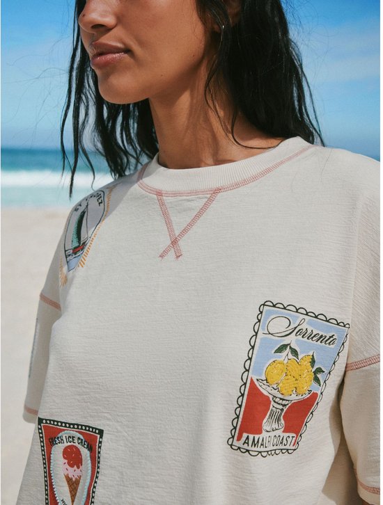Vrouw draagt een crème T-shirt met 'Sorrento' en 'Amalfi Coast' emblemen op het strand.