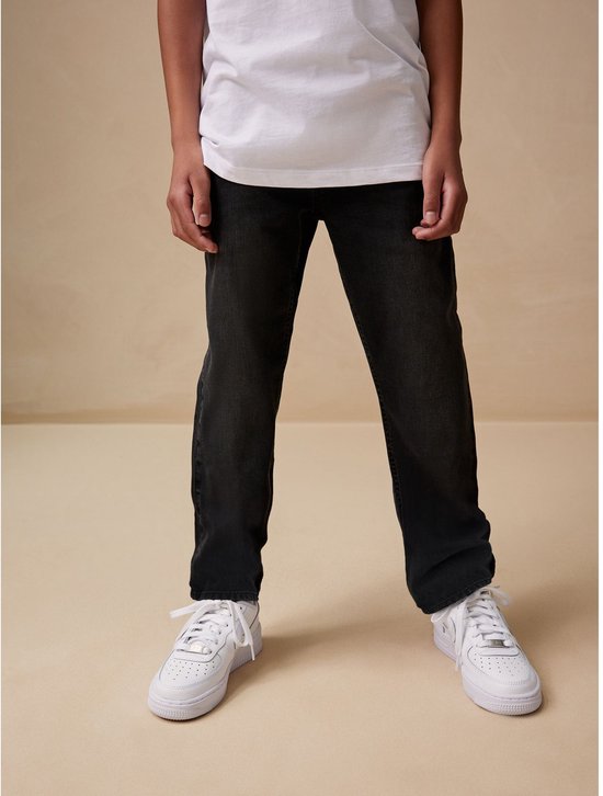 Jongen in een witte T-shirt en zwarte spijkerbroek, staand, met witte sneakers.