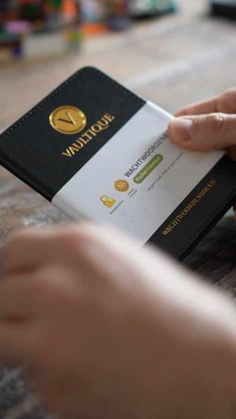 Een hand houdt een zwart wachtwoordenboekje met gouden logo van Vaultique vast, met een papieren band met informatie.