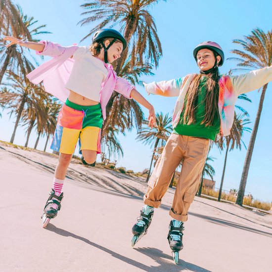 Twee meisjes met helmen op rolschaatsen bij palmbomen, in kleurrijke kleding.
