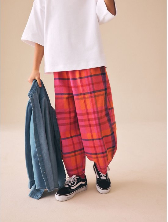 Kind in een witte top en wijde, geruite broek in roze, oranje en rood.