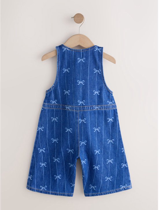 Achterkant van een blauwe denim jumpsuit zonder mouwen met strikjesprint, hangend aan een houten kledinghanger.