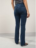 Model draagt een donkerblauwe spijkerbroek met bootcut pasvorm en een slim-fit model. De broek sluit hoog op de taille en heeft een metalen detail op de achterzak.