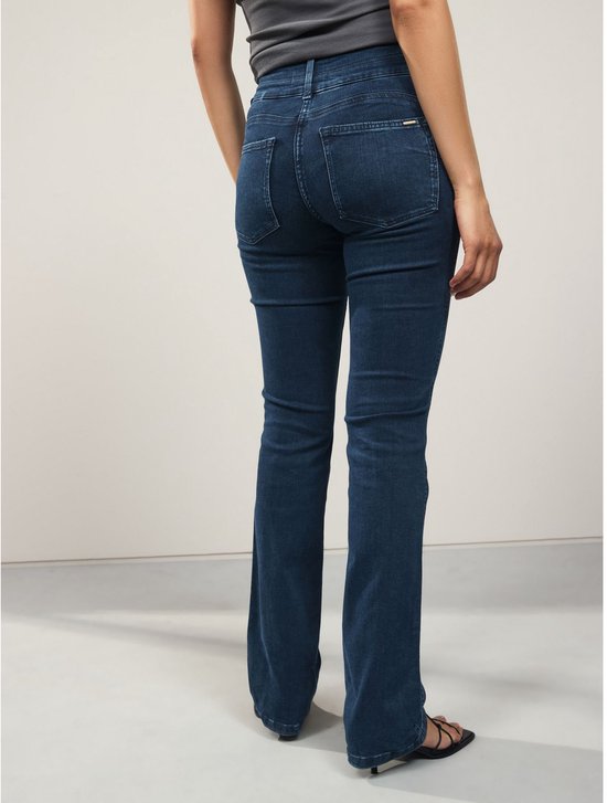 Model draagt een donkerblauwe spijkerbroek met bootcut pasvorm en een slim-fit model. De broek sluit hoog op de taille en heeft een metalen detail op de achterzak.