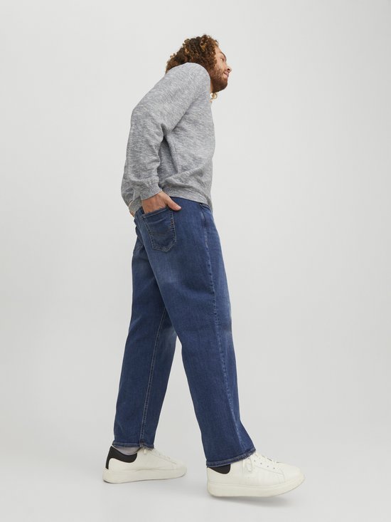 Man met krullend haar, van opzij gezien, draagt een lichtgrijs gemêleerde sweater en een spijkerbroek van blauw denim.