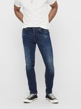 Man met wit T-shirt en donkerblauwe jeans, staand, met handen in de zakken.