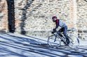 Man fietst op een racefiets in een stad, draagt een zwarte jas en een fietsbroek.