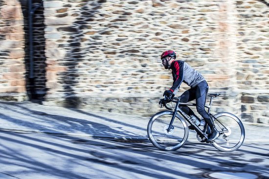 Man fietst op een racefiets in een stad, draagt een zwarte jas en een fietsbroek.