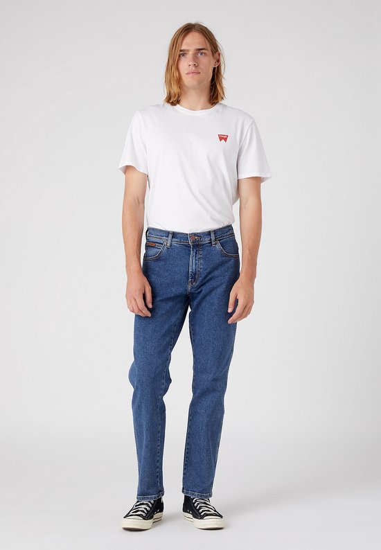 Man met lang blond haar, staand, in blauwe spijkerbroek en wit t-shirt met logo.