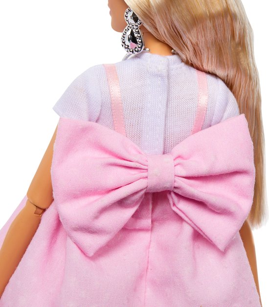 Barbiepop met grote roze strik op de rug van de jurk, een witte top en sprankelende oorbellen.