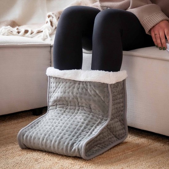 Grijze voetenwarmer met witte pluche voering, gedragen door een persoon op een bank.