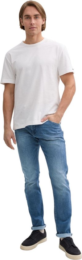 Man met hand in zak, draagt witte T-shirt en blauwe jeans.