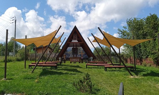 Geelbruine schaduwdoeken boven een kapel en zitbanken in een grasrijke omgeving.