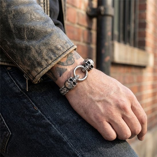 Zilverkleurige schakelarmband met schedelmotief, gedragen aan een arm naast een bakstenen muur.