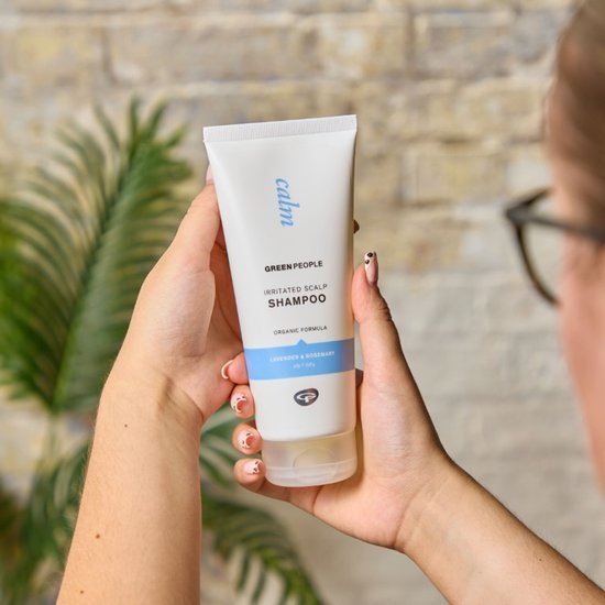 Handen houden een witte tube met 'calm' erop in blauw, 'Green People Irritated Scalp Shampoo', met blauwe band en lavendel & rozemarijn tekst.
