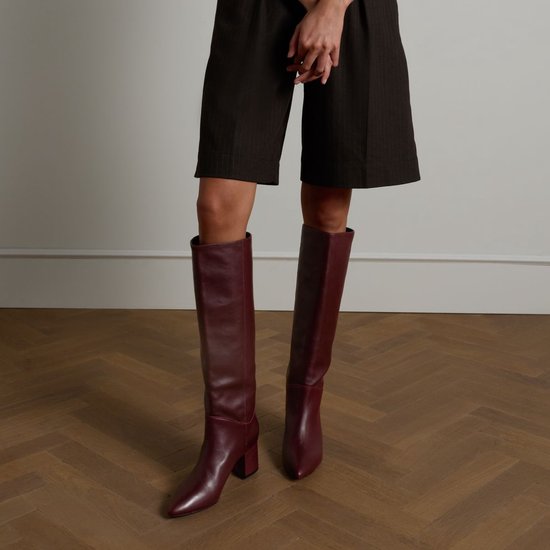 Bottes hautes en cuir bordeaux à bout pointu et talon bloc, portées par un modèle sur un plancher en bois.